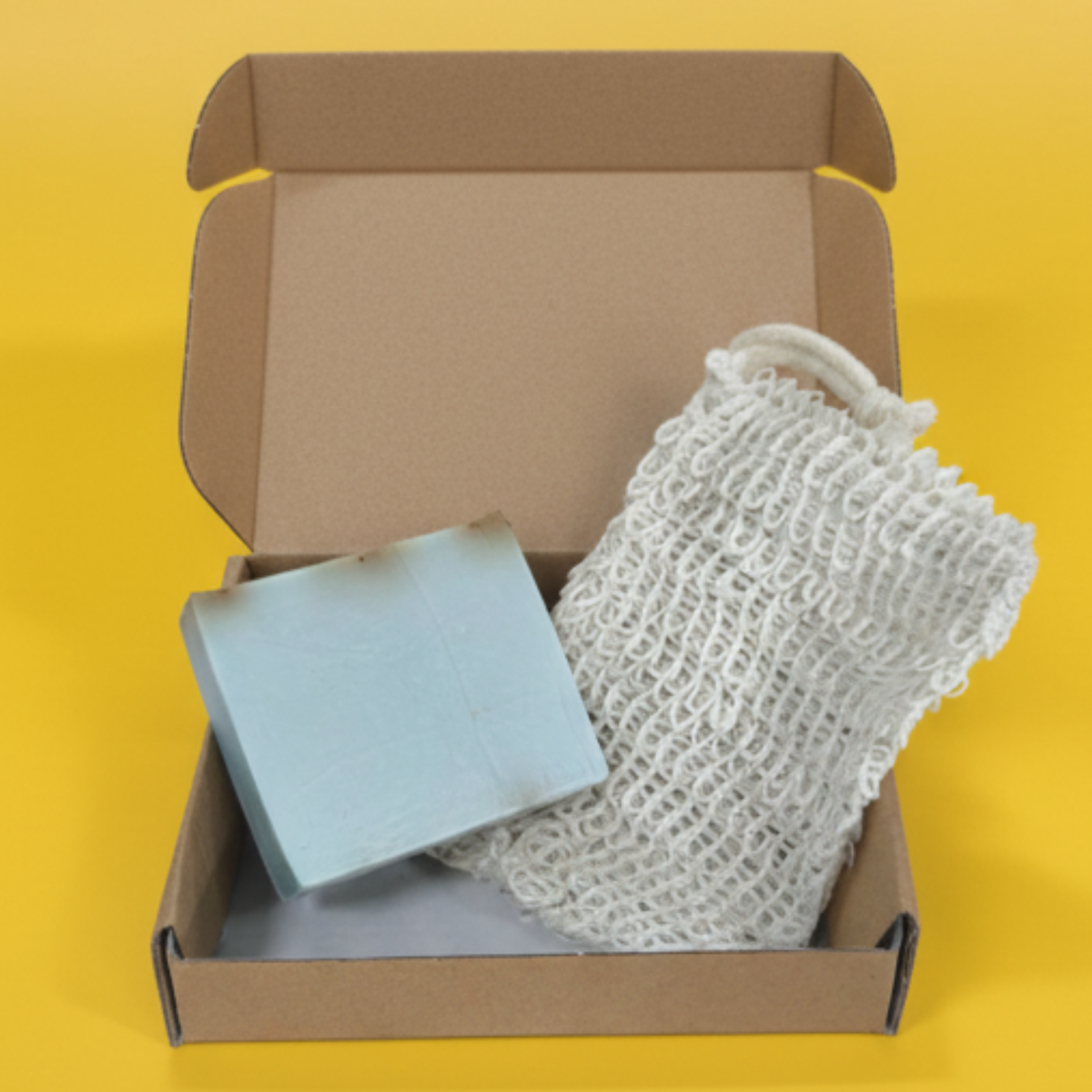 An open cardboard box containing a white mesh bag and a small rectangular object, possibly a soap bar, against a yellow background.