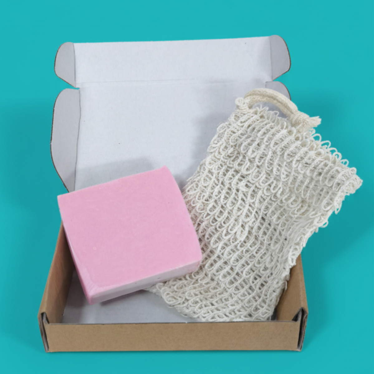 An open cardboard box containing a pink bar of soap and a white mesh bag, placed against a teal background.